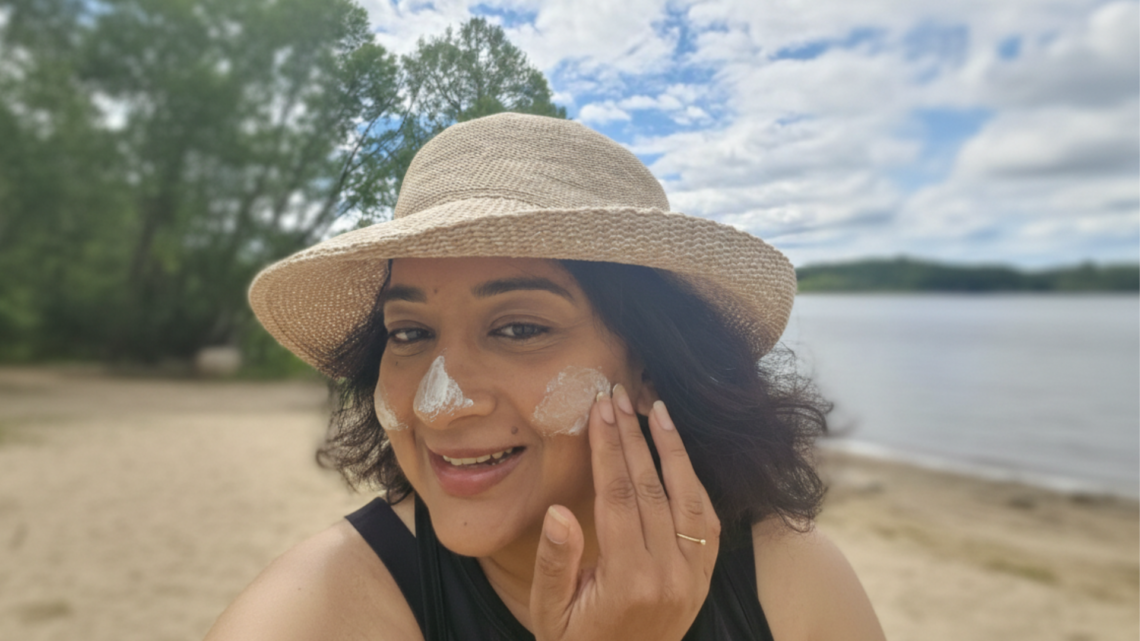 Indian-Australian woman applying mineral sunscreen to her face outdoors by a lake, demonstrating real-life wear on medium-deep brown skin.